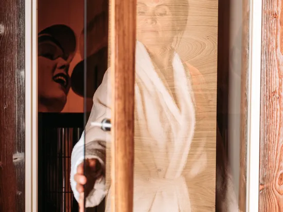 Woman in white bathrobe opening glass door of wooden infrared sauna